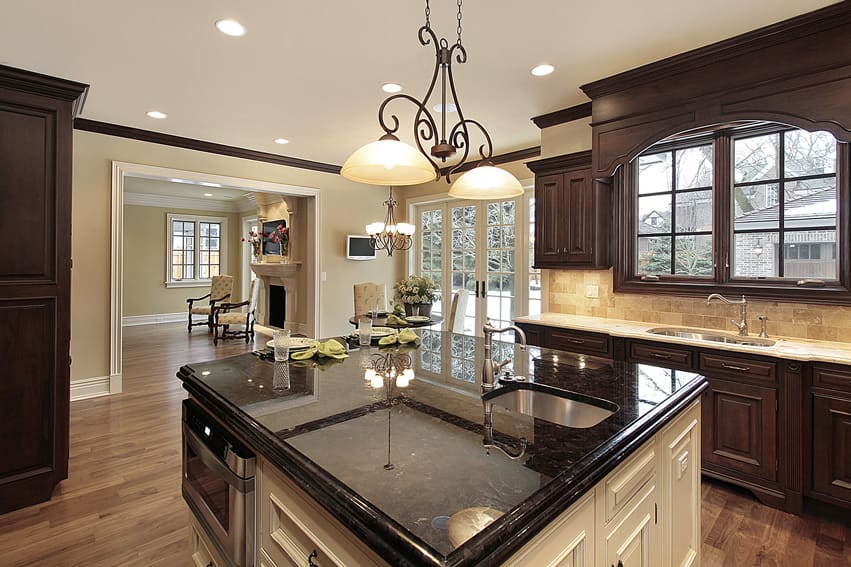 This kitchen reflects modern interior design ideas with brilliant wooden floor and light cream walls and somewhat counteractive yet brilliant traditional cabinets. The dark walnut wood-made cabinets pop with granite top against a backsplash of sandstone tiles. The cabinet of the large island is white with blue pearl granite countertop and placed beneath low hanging lights.
