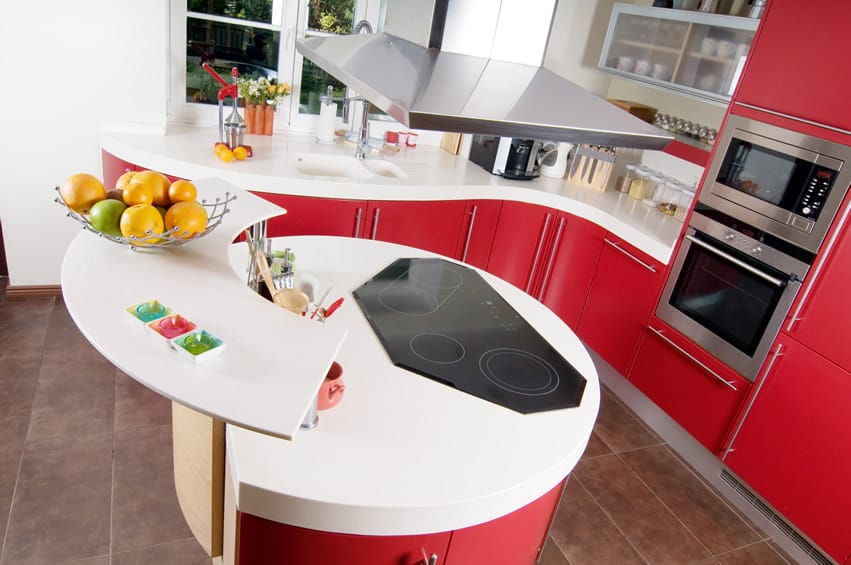 A cute modern kitchen with red finished cabinets and white countertop. The stainless steel oven , handles and vent hood adds a shiny feel . The circular shape of the island looks great too.