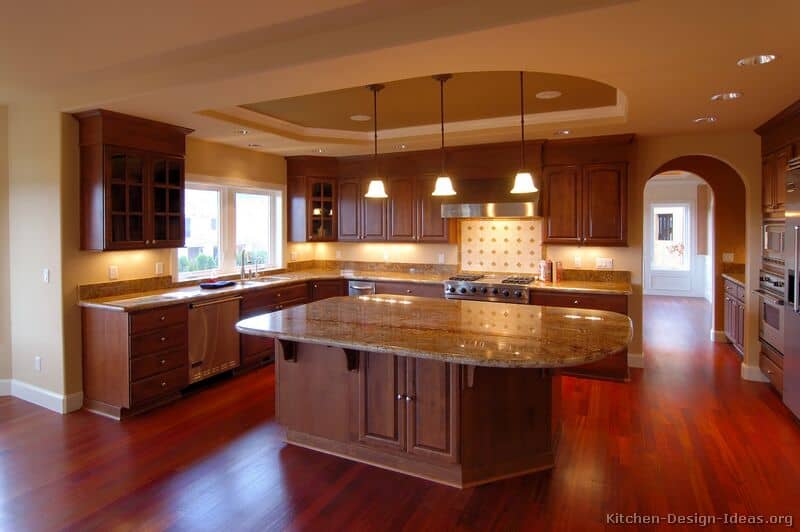 This contemporary kitchen features a reddish glow. The floor is red with cherry wood finishing. The brown wooden cabinet and island is covered on top with extended marble countertop. The low hanging lights and low spotlights below the cabinets lit up the room.