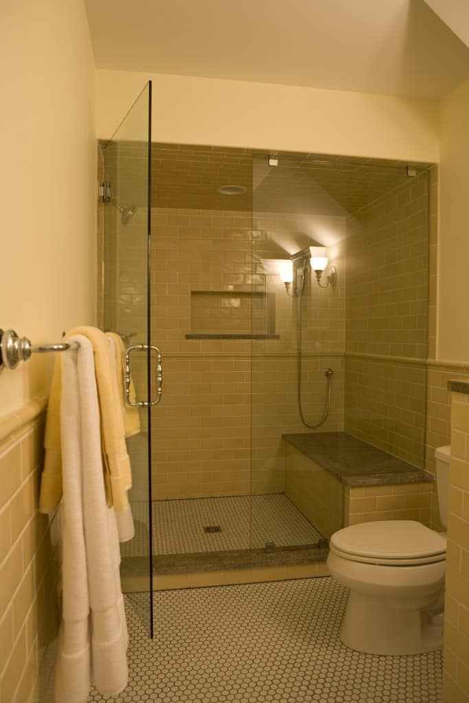 A small bathroom with hexagonal tiles used for flooring . The wall has brick textures with beige color. The main shower area is kept separated by glass partition and has a granite colored seat