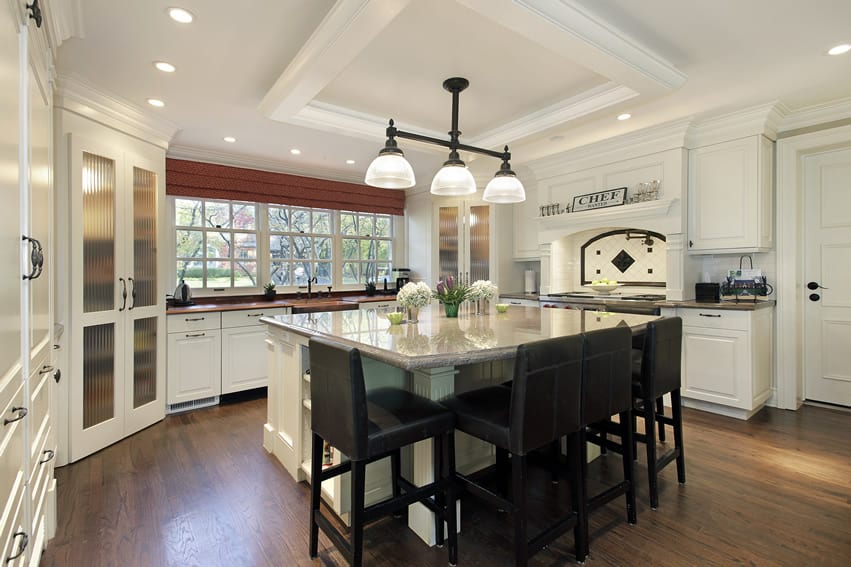 This kitchen has neo-classic style cabinets with white finish. The white coloring on the wall and island make the grains of the walnut wooden floor pop. The dark stools with brown upholstery adds to the contrasting color combinations.
