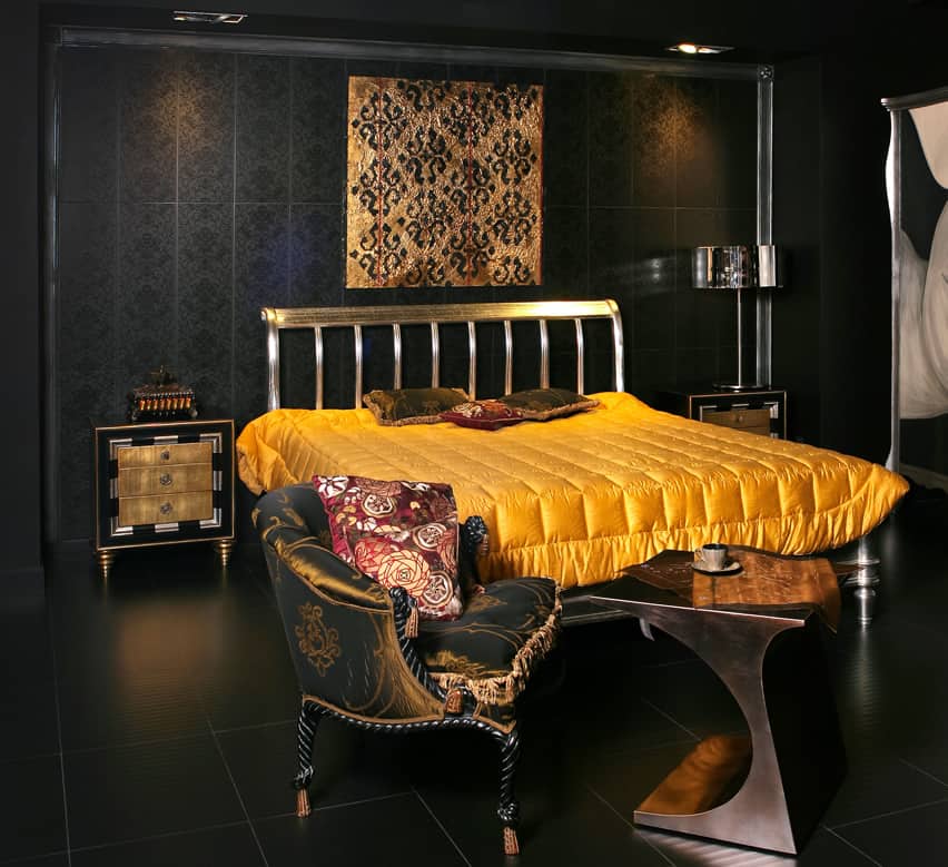 Ultra-modern, minimalist and masculine bedroom. Flooring, walls and ceilings- all are black. There are lots of oriental vibe with golden bedside tables, gold-stained metal in bed frame, yellow bed sheet and red floral pillows.
