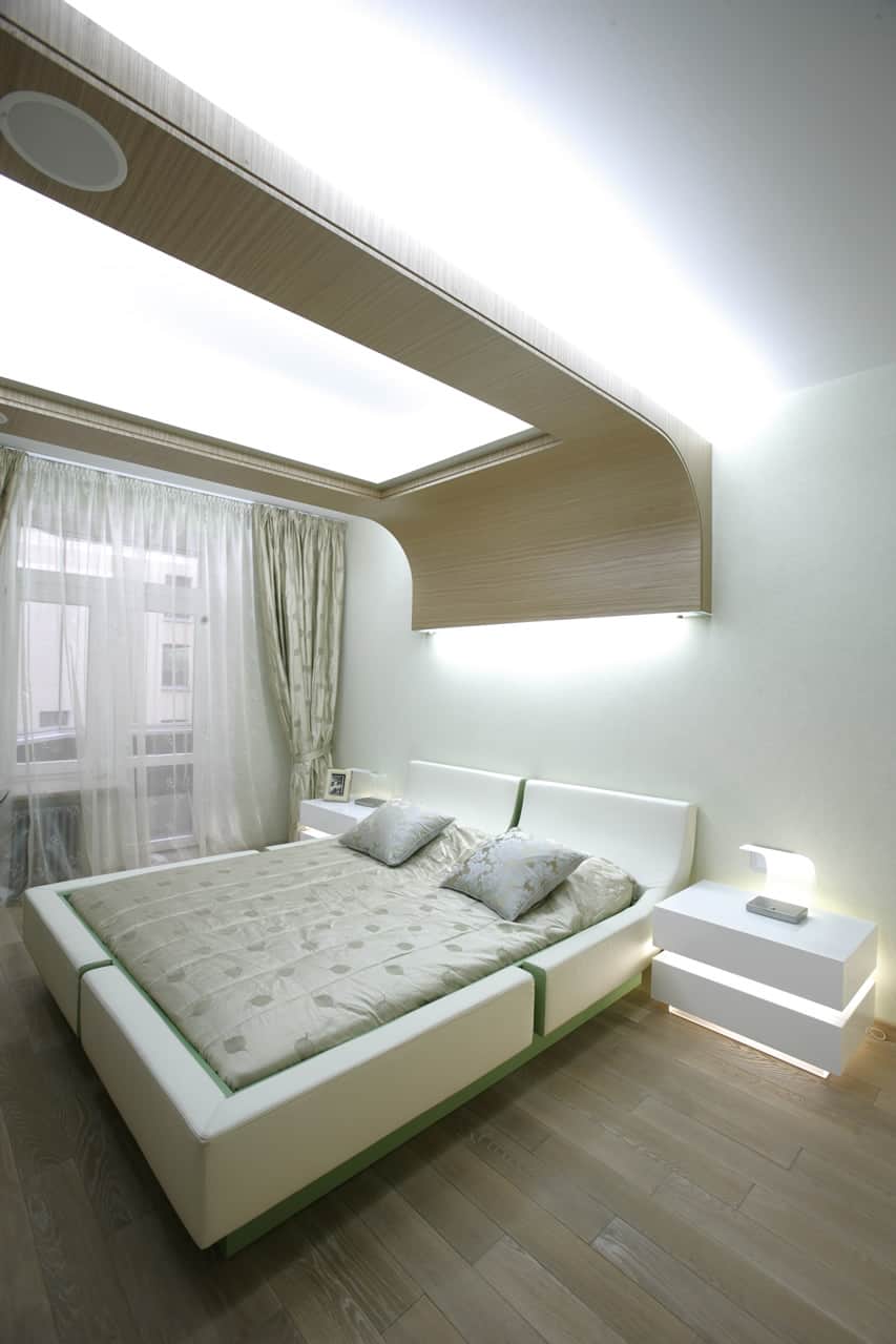 An ultra-modern bedroom that uses futuristic forms . The white and green bed has a curved design . The large portion hanging over the bed has a curved canopy with maple laminates. The flooring is made of wood . The spot lights placed near the head and the side table laminates the room even more.