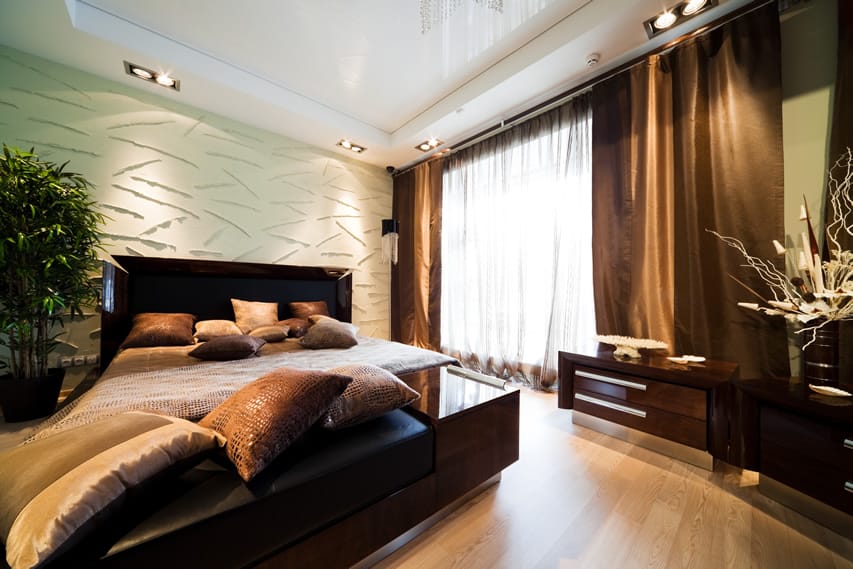 A state-of-the-art bedroom that makes use of a variety of textures . The accent wall has a 3D wall panel while brown color dominates the room. The bed uses dark wood with black upholstery . The large potted plats adds some greenery into the room.