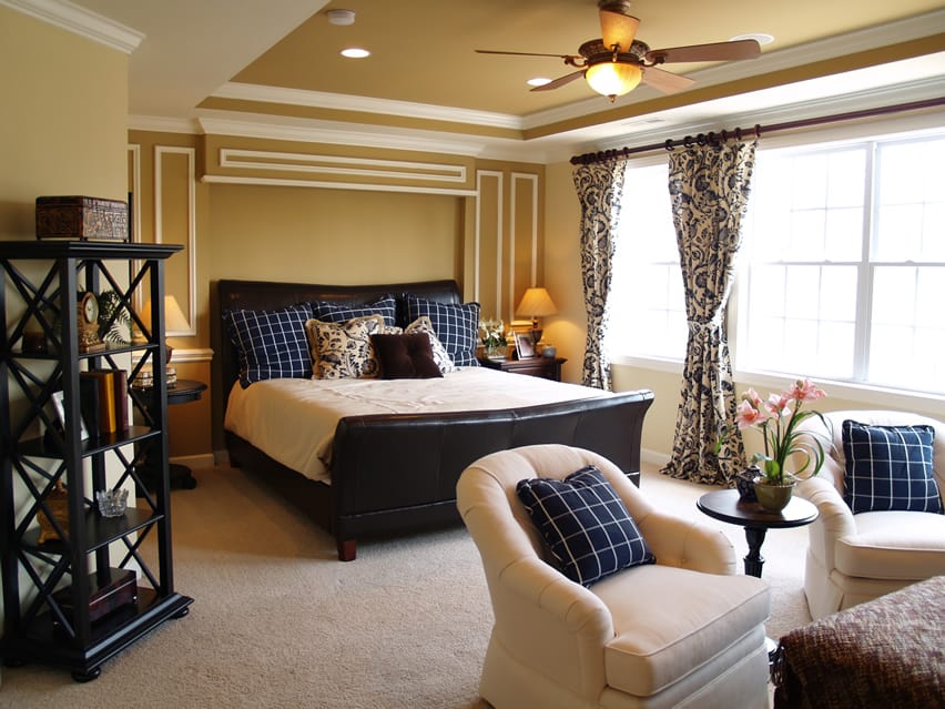 A classic bedroom that uses gold accent walls that are fitted with white stripes. The bed sheet and sofa are light colored and so too is the carpet. The black bed frames pop against the light colors. White checkered fabrics on blue color is used in the cushions on bed and sofa. The lamp shades have a beautiful orange glow.