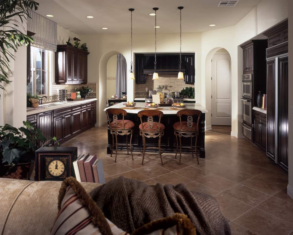 Dark marble flooring and dark wooden cabinets make this kitchen impressive . The dark island with white marble countertop has several ornate seating arrangement. The dark genre of the kitchen is contrasted by white walls and the arched doorways.