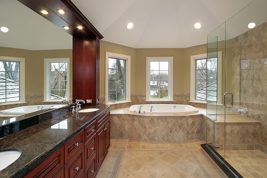 A charming bathroom that features red cherry wood vanity with dark countertop and a large mirror with spot lights used overhead. The roof is prism shaped on the far corner and the far side wall contains two beautiful diagonal windows.