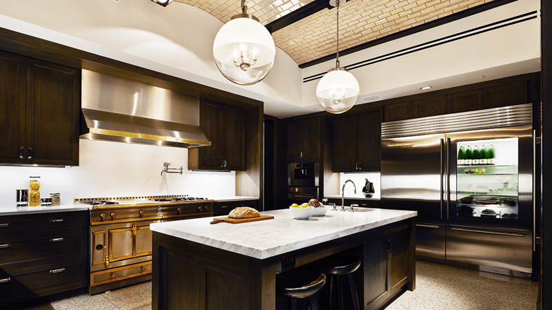 A large L-shaped modern kitchen that features a sharp contrast between dark and light colors. Large globe-shaped lighting and white countertops enlightens the room .
