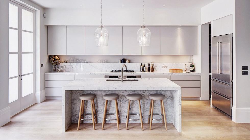 An all-white kitchen with white cabinetry and central marble-made island having seating arrangements. The flooring is made of light woods.
