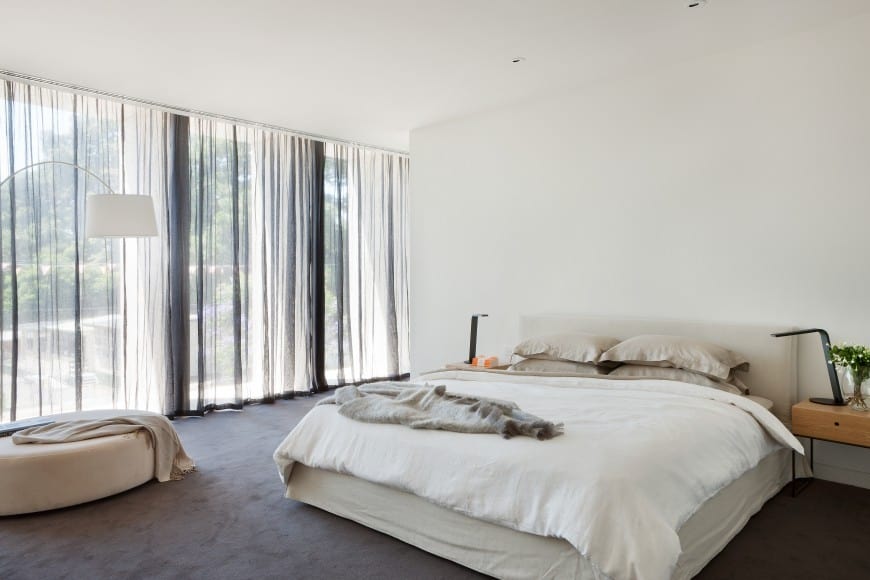 A modern bedroom with white walls and roof. Large window allows a lot of light to come in. The whole floor is covered with grey carpet . A peaceful and soothing vibe is generated from the placement of organic shapes and textures.