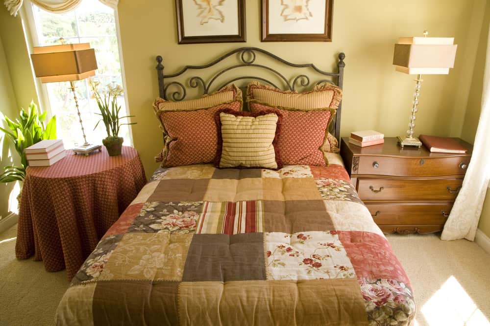 A country style bedroom with blanket full of square patterns of different colors. A wooden cabinet is placed on one side while a round table is placed on the other side. The glass square lamps and green potted plants complete the decorations.