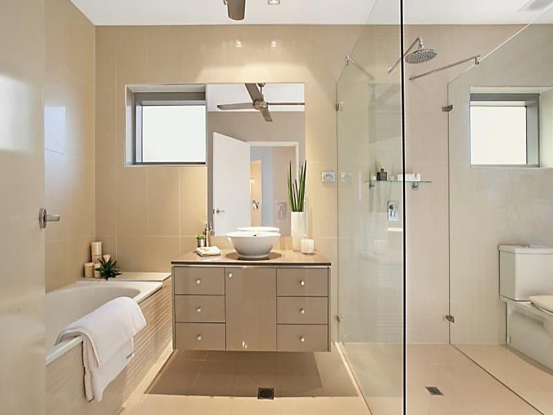 Light brown colored wall tiles and floor finishes are matched by the horizontal textured bathtub outer wall of the same color. Two glass partitions separate the shower area and the bathroom.