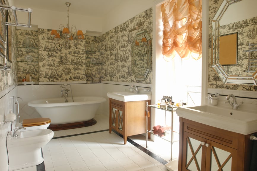 A white bathroom with nice wallpapers. Two vanities are placed by the sides of the large window. The toilet seats are placed on the opposite side of the vanities.