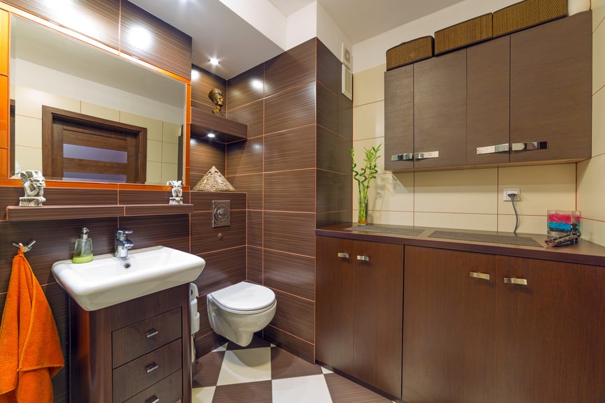 An elegant bathroom with dark wood cabinetry and dark brown tiles . The mirror is bordered with beautiful orange frame.