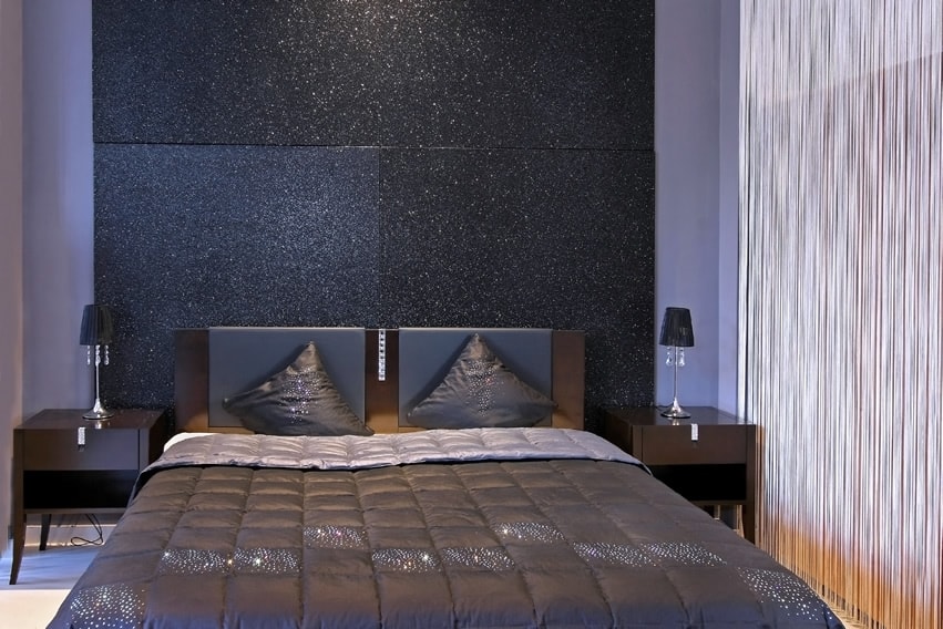A glamorous small bedroom with blue walls . The head side wall hosts a square glittery black panel. The bed frame and table are deep colored while the headboard has deep grey leather upholstery. The bed sheet matches the overall atmosphere rather spectacularly.