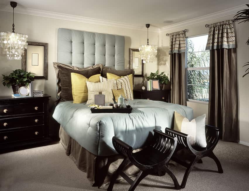 A dark, moody yet delightful master bedroom that uses a splendid mix of contemporary and classic pieces. The furniture and frames are dark colored placed over a grey carpet. The headboard is catchy with cushy light blue while the pillows are mainly yellow and cream colored.