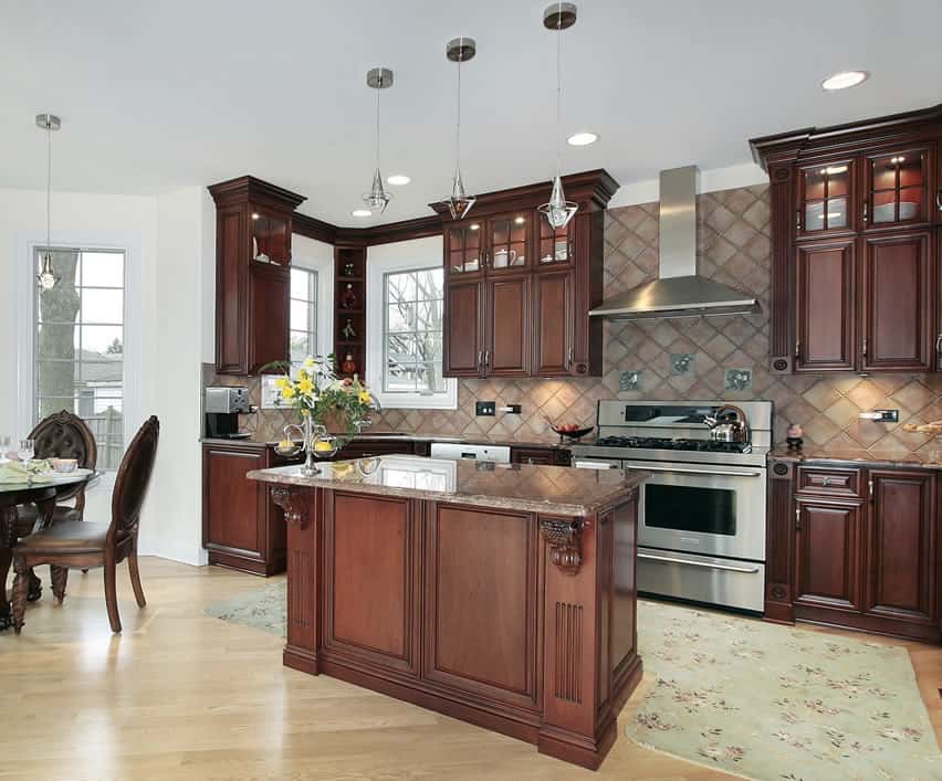 The light floor made of oak wood and plain white walls make the dark cherry cabinets and the island pop even more. The reddish stone backsplash looks awesome behind the cabinets and silver- colored appliances. The artistic  island is small but novel.
