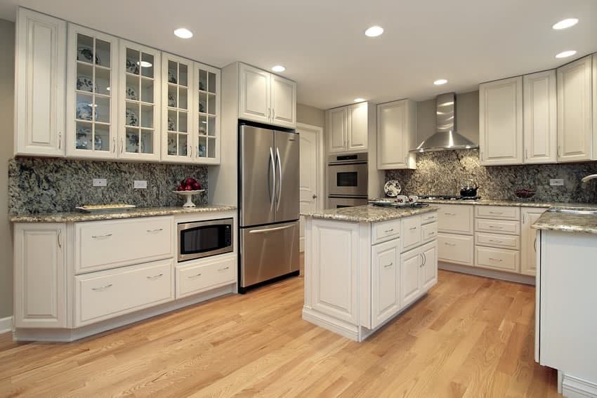 This kitchen uses neutral colors with oak wooden floor and gray wall paints. The extended gray granite countertops goes on rather well with the neutral background & white cabinets and island. A section of the overhead cabinet near the refrigerator uses glass doors with dishes inside.